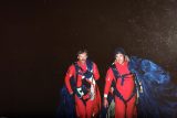Two skydivers in red suits walk together in the dark, carrying blue parachutes. They appear satisfied and are equipped with various gear.