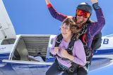 Two people tandem skydiving, exiting a plane mid-air. The instructor wears a helmet and sunglasses, guiding the other person who is smiling with arms raised. The plane is white and blue with an open side door. Clear blue sky in the background.