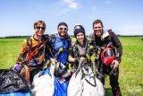 Four skydivers stand smiling on a grassy field, wearing their parachuting gear. They hold their parachutes in hand, and the sky is clear and sunny.
