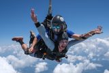 Two people are tandem skydiving against a backdrop of a clear blue sky and fluffy white clouds. The person in front is smiling and has arms outstretched, while the instructor is behind, guiding the descent. Both are wearing helmets and safety gear.