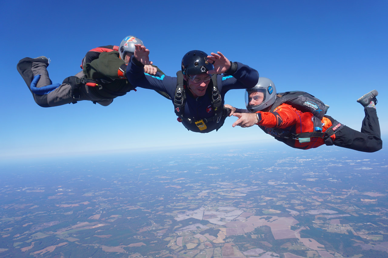 Three skydivers in mid-air formation, wearing jumpsuits and helmets, against a backdrop of clear blue sky and terrain far below. The central skydiver is reaching out, with the others extending towards him, showcasing teamwork and coordination.