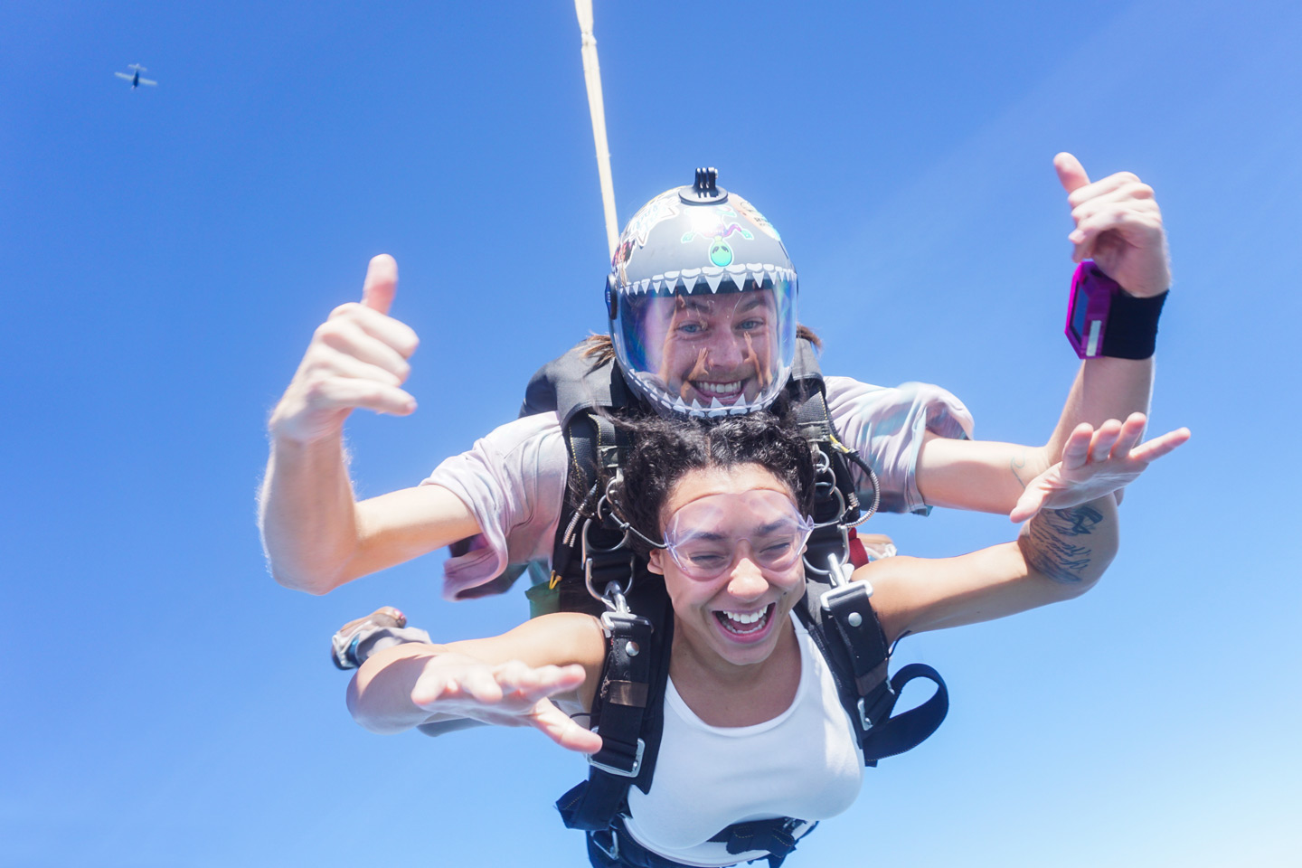 A tandem skydive in progress, showing an instructor and a smiling woman free-falling against a clear blue sky. The instructor gives a thumbs-up, and both wear skydiving gear. A small plane is visible in the distant sky.