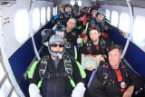 A group of people wearing skydiving gear and helmets are seated inside an airplane. They are preparing for a skydive, with parachutes visible on their backs. The interior of the plane has blue and white walls, and there are windows along the sides.