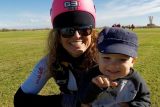 A woman wearing a pink helmet and sunglasses is smiling while holding a small child in her arms. They are outdoors on a grassy field under a blue sky with some clouds. The child is wearing a dark baseball cap and smiling.