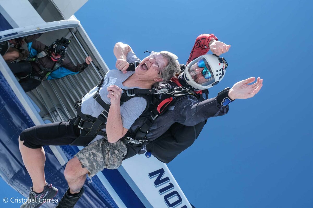 Older woman with short gray hair exiting the airplane on a tandem jump with an instructor in a while helmet.